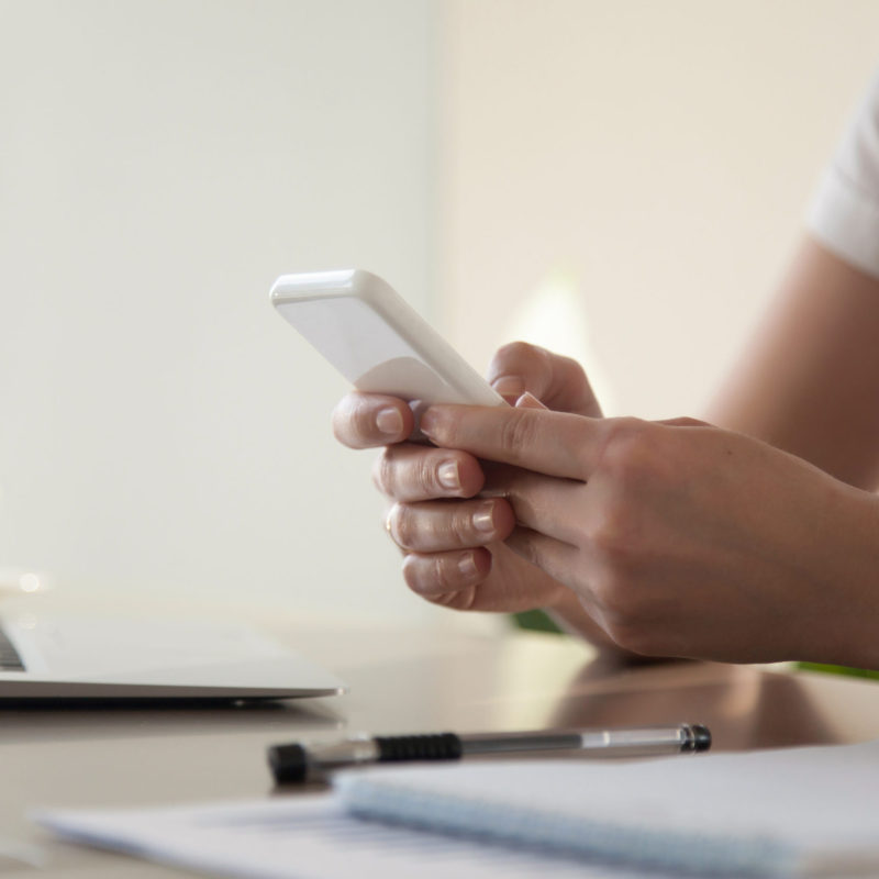 Close up image of modern cellphone in womans hand at desk with laptop. Office worker boring at workplace and playing games, writing in social networks. Businesswoman looking important contact on phone